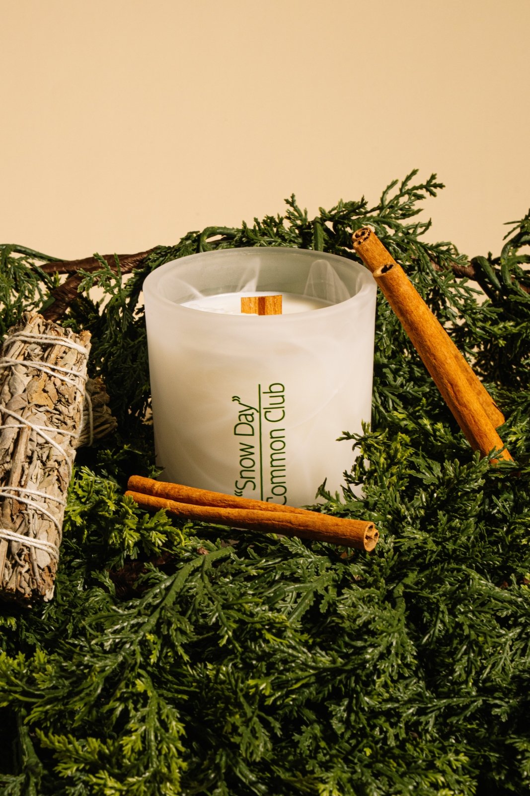Candle with 'Snow Day Common Club' branding on a bed of greenery with cinnamon sticks and sage.