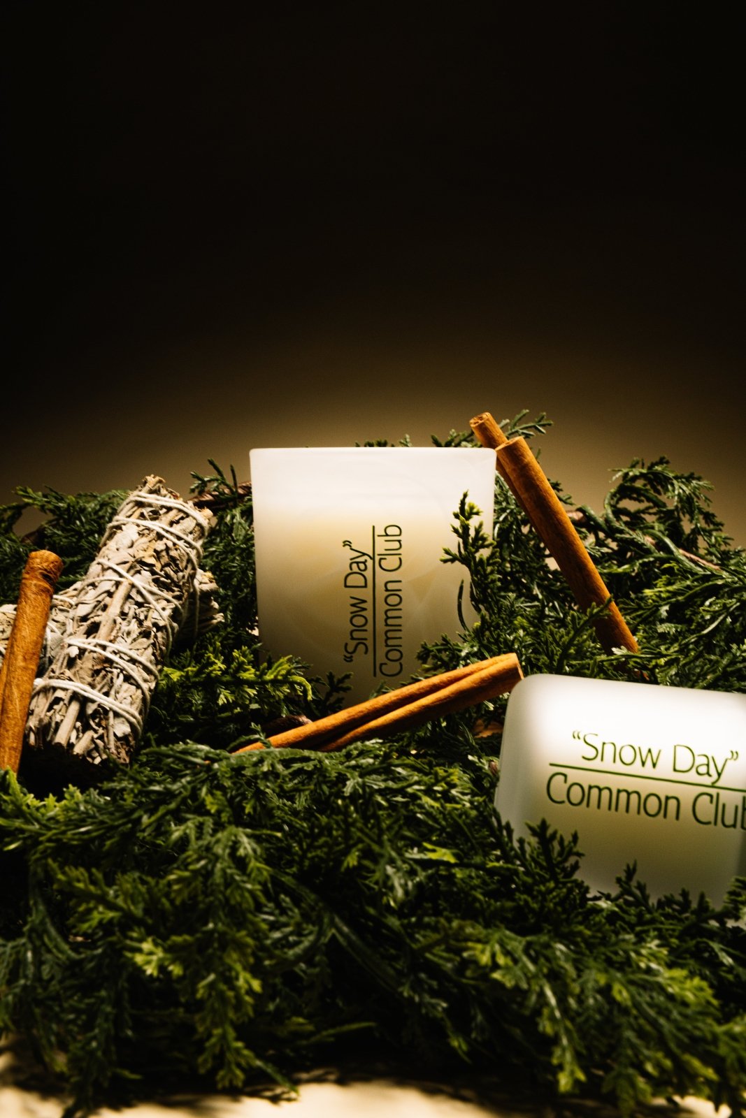Two candles labeled 'Snow Day Common Club' on a bed of greenery with smudge sticks.